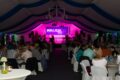 An illuminated marquee where many people sit at tables and look towards the stage, above which hangs a banner with the inscription 