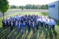 Gruppenfoto einer Firmenbelegschaft, die auf dem Rasen neben einer Produktionshalle steht.