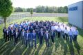 Group photo of a company workforce standing on the lawn next to a production hall 